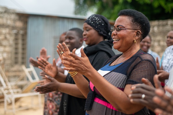 In Kenya, a woman with cropped hair, glasses and a colorblock dress stands at forefront of a line of five women, singing and clapping. They are members of an activist collective.