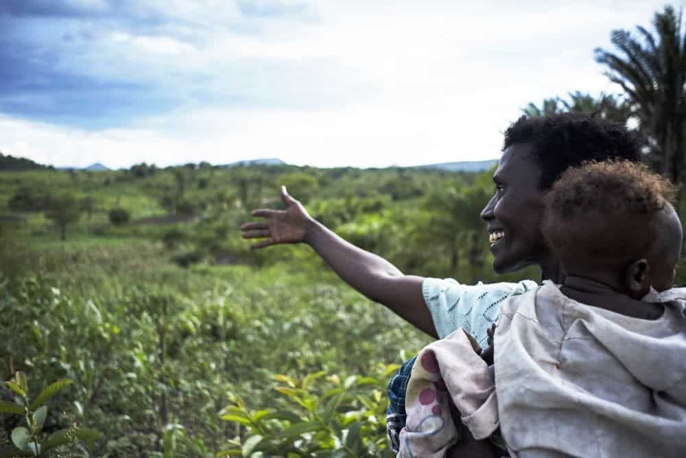 A mother carries her child and gestures toward a lush field in a region threatened by oil extraction