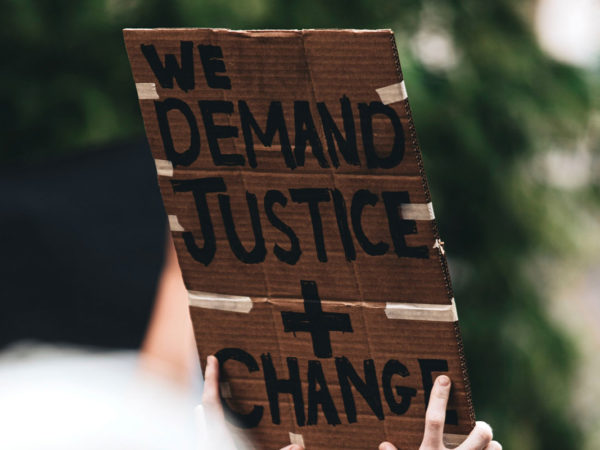 At A Protest, A Sign Written On Cardboard Reads, "We Demand Justice + Change."