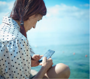 This image shows a woman from behind, as she looks down at her phone while sitting near the sea. She helps migrants who are stuck at sea, and example of how we can support activists,
