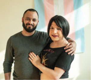 A man with a beard has his arm around a trans woman standing in front of a flag in support of LGBTQ rights. Support activists like these today.