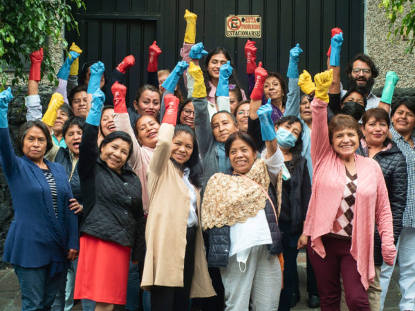 A Group Of Domestic Workers In Mexico Stand Together, Wearing Colorful Dishwashing Gloves, Arms Raised In Celebration.