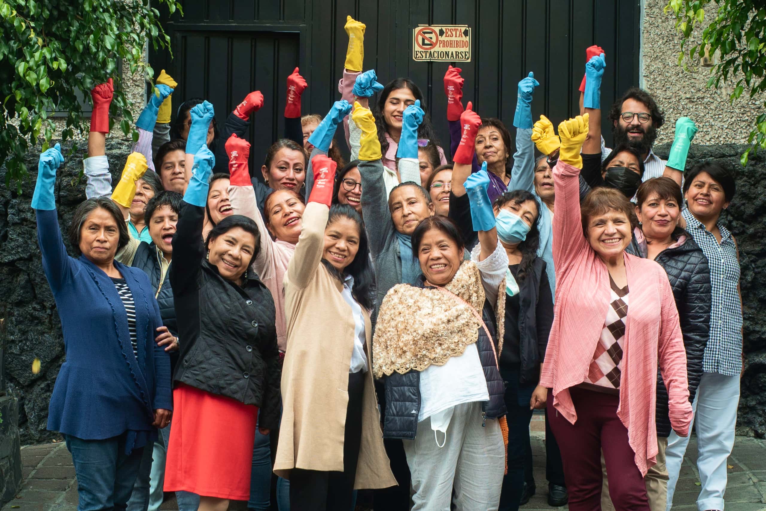A group of domestic workers in Mexico stand together, wearing colorful dishwashing gloves, arms raised in celebration.