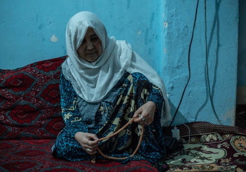 Woman wearing a colourful dress and hijab holds her beaded necklace in a dim blue room.