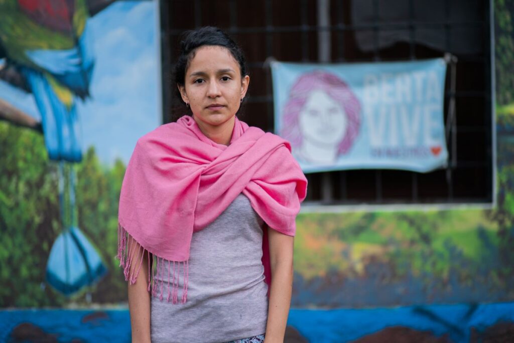 A young woman wearing a solemn expression and pink scarf stands in front of a poster memorializing her mother, a murdered activist.