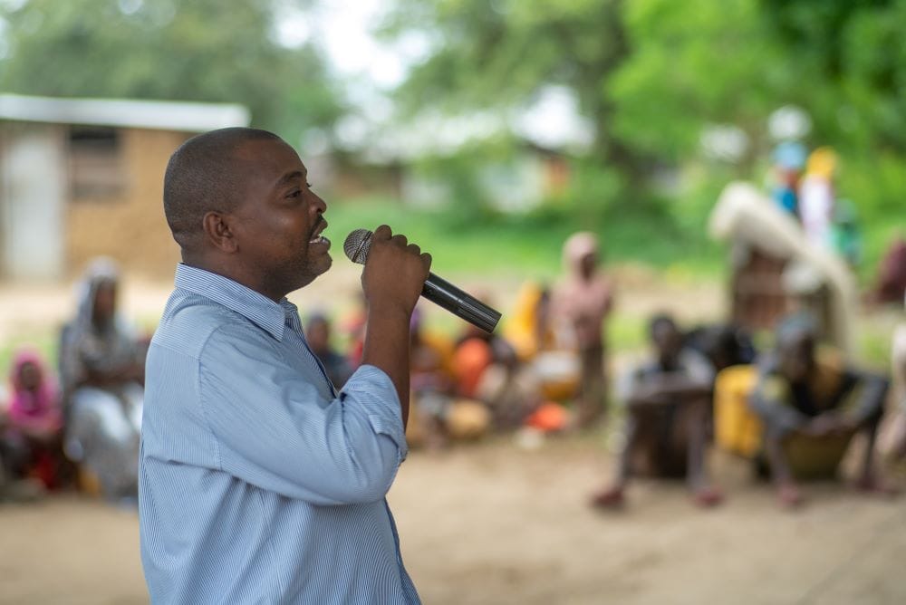 A Kenyan man in a blue shirt holding a microphone addresses a small group seated outside