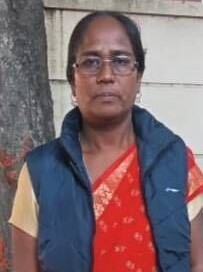 A woman wearing a red and gold sari with a blue vest and glasses and a solemn expression