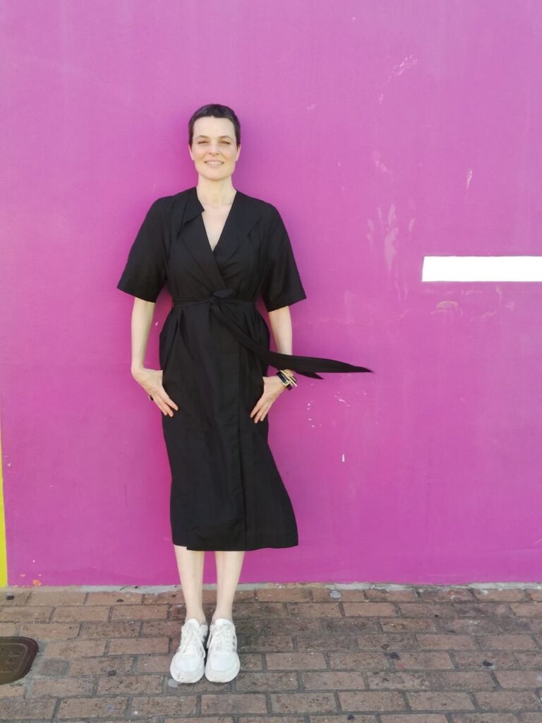 A person in a black dress stands in front of a pink wall.