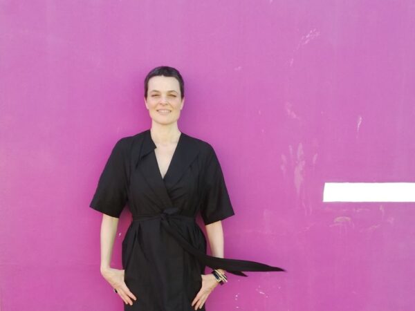 A Person In A Black Dress Stands In Front Of A Pink Wall.