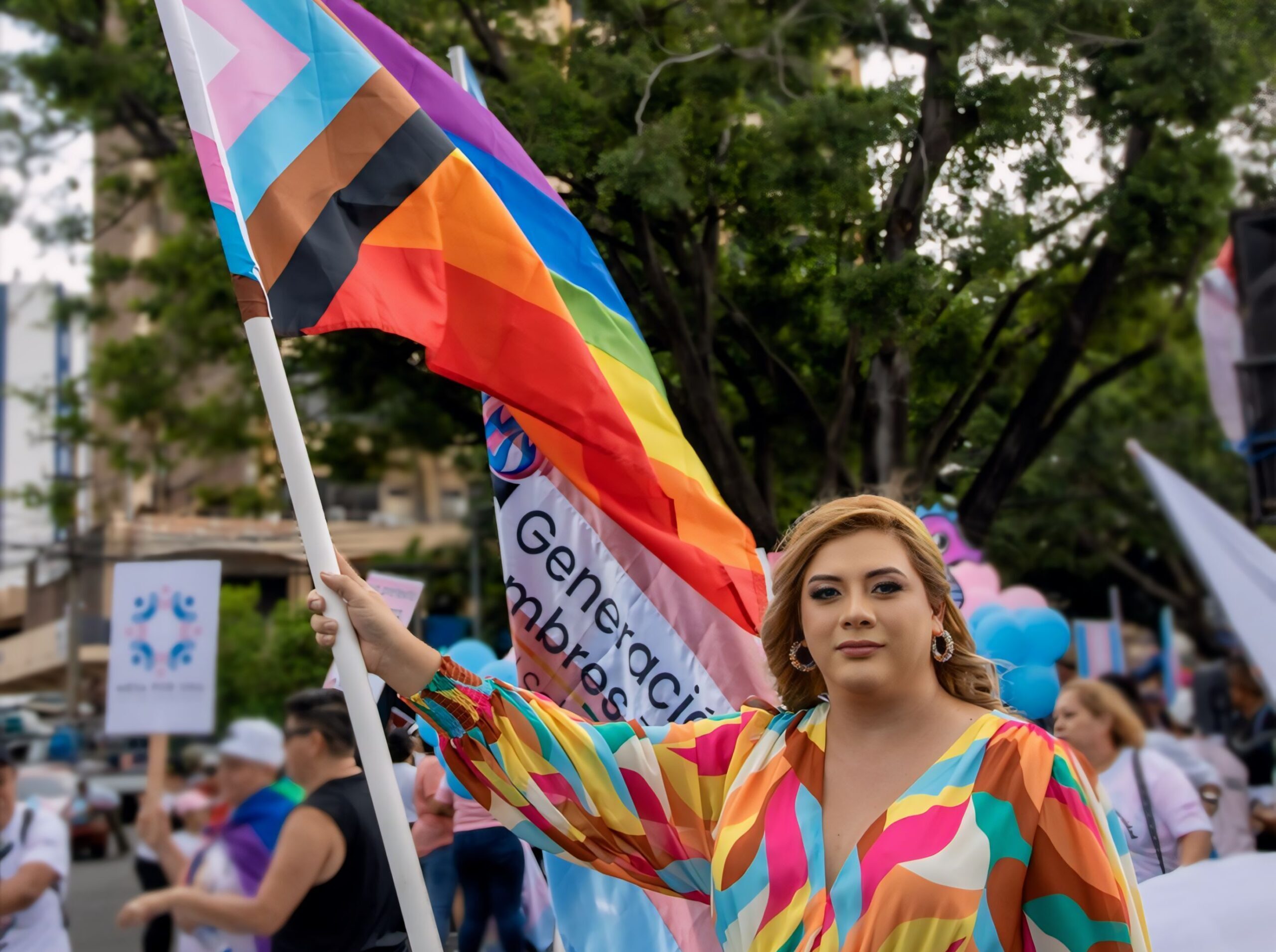 A trans woman wearing a multi-colored dress walks during a Pride parade, the progress pride flag in her right hand.