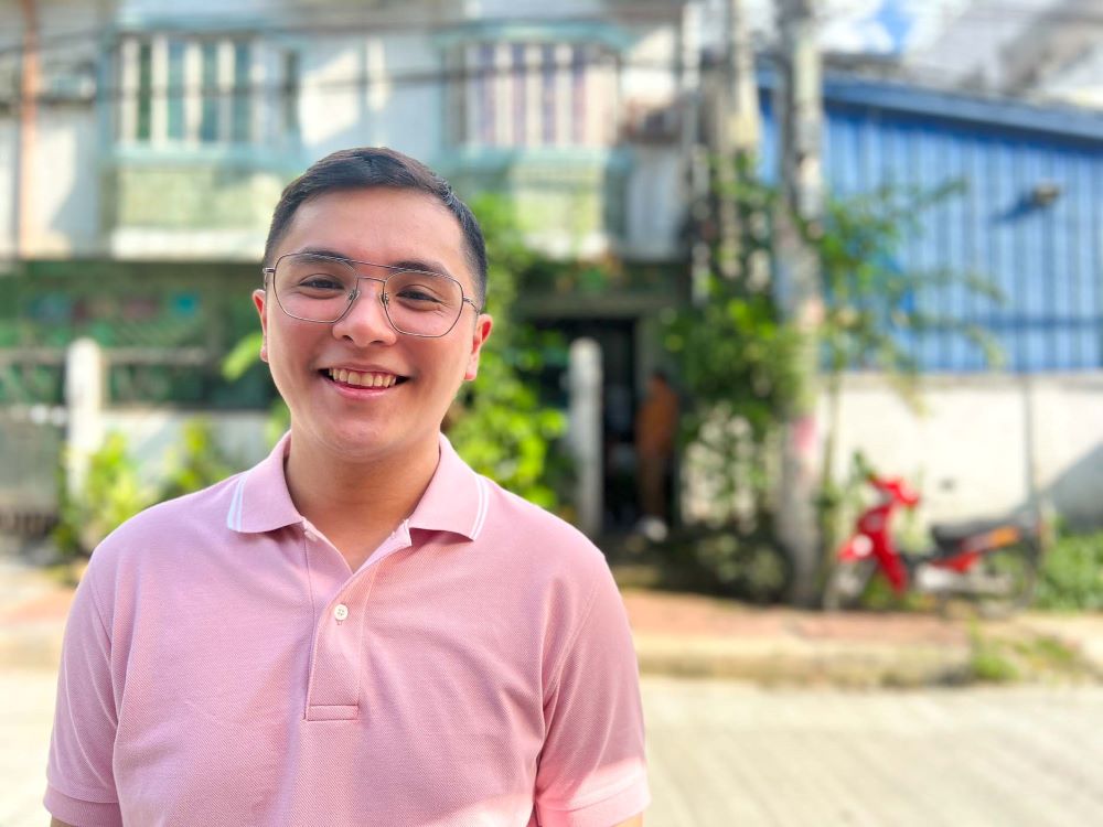 A young Filipino man with glasses wearing a pink polo shirt stands in front of a university building.