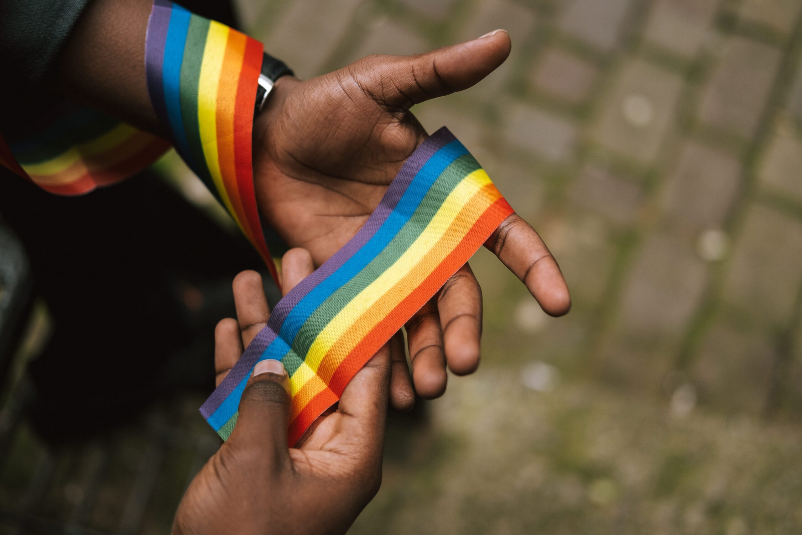 A Black man holds a rainbow ribbon in his hands. His face is not shown,